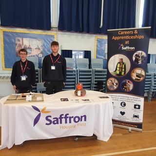 Two Saffron Apprentices Standing Behind Marketing Table 